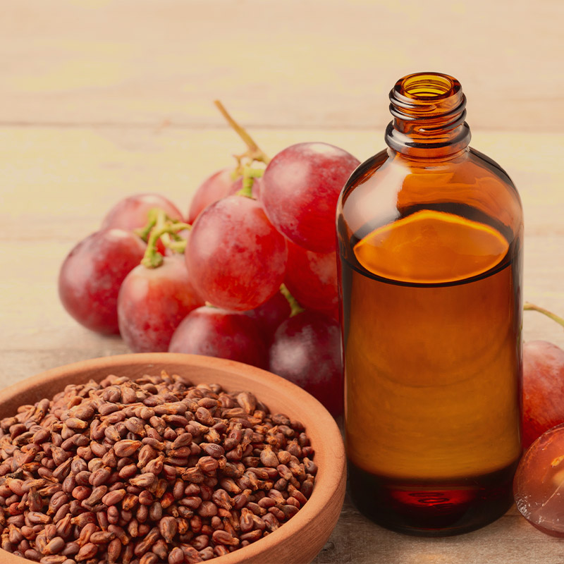 Guapo's Natural Ingredient: A wooden table displays a bottle of grapeseed oil alongside a cluster of grapes, the source of the oil. Tiny grapeseed seeds are scattered on a clay tray nearby.