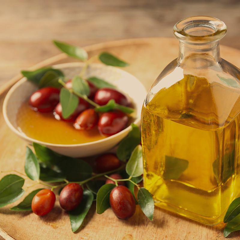 A bottle of jojoba oil sits beside a bowl containing the oil and jojoba seeds, the source. Jojoba leaves complete the display, showcasing the natural ingredients in Guapo's products.